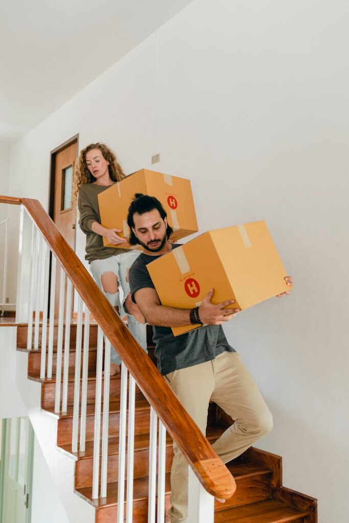 pexels photo 4246097 Concentrated couple in casual clothes carrying carton boxes with personal items down stairs while moving to new apartment