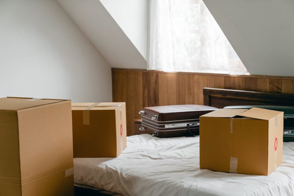 pexels photo 4246100 Small cardboard boxes and leather cases placed on bed with sheet near big box in light room of house under sloping roof